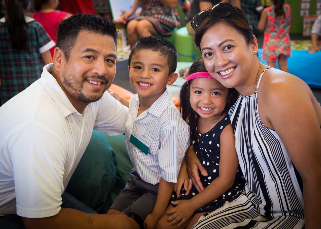 A young family, smiling family sitting together on the floor at a gathering. The father is on the left, mother on the right, with a young boy and girl (under 6 years of age) sitting between them.
