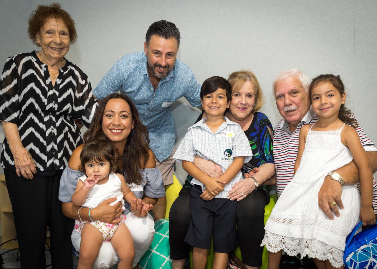 Three generations of family sit and stand together, smiling for a family photo. Mum, dad, grandfather, two grandmothers and three children are posing for the photo.
