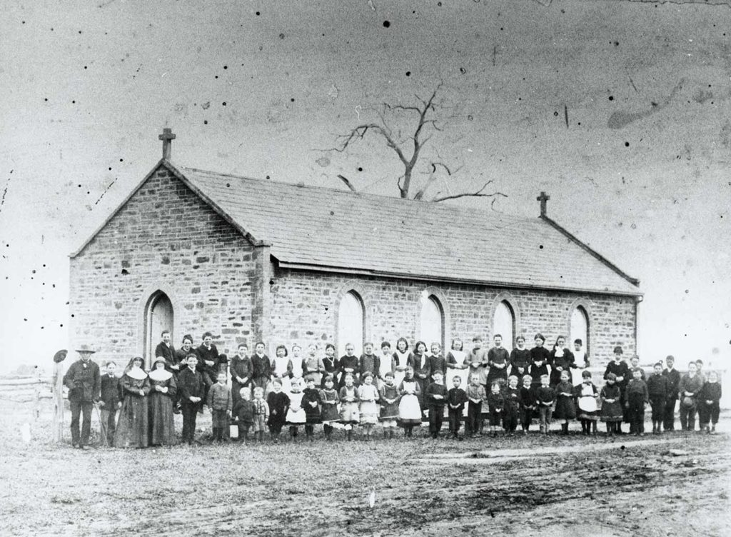 A historical, black and white photograph of an early Australian school in the bush. All of the students are standing in a line out the front of the single building.